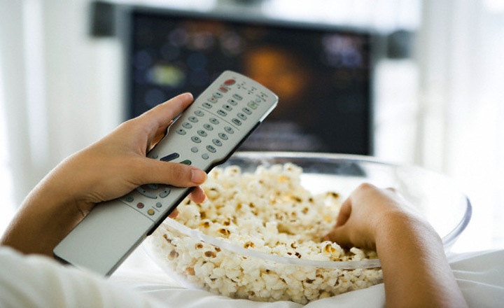 Person watching television, holding remote control and bowl of popcorn, cropped view